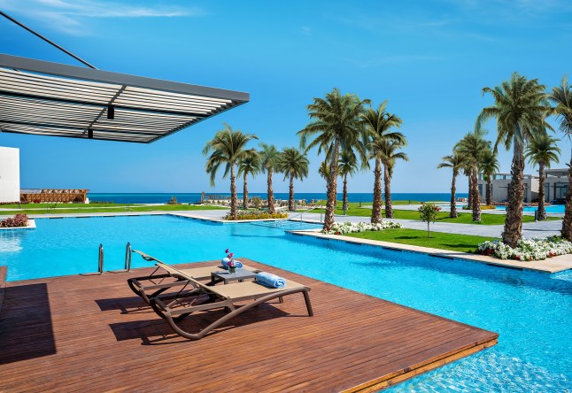 A view of a dazzling blue pool surrounded by grassy patches and palm trees at the Rixos in Hurghada. The Red Sea can be seen in the distance.