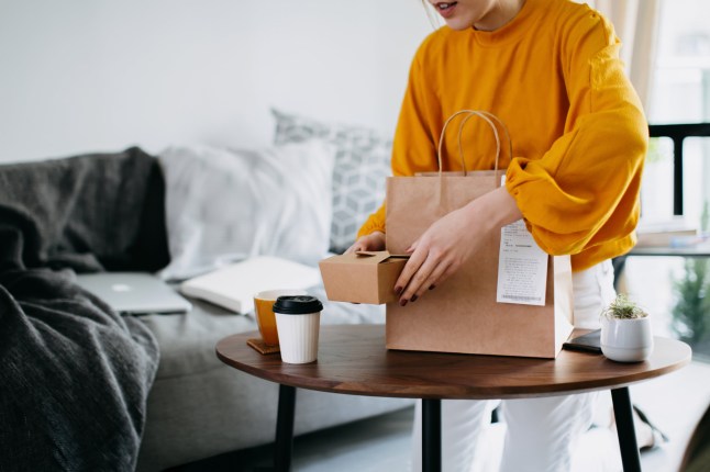 Cropped shot of young Asian woman holding boxes of home delivery takeaway food that have just arrived to share, serving on the coffee table. Eating at home concept